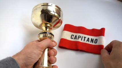 Man's hands hold football captain's armband and cup won when he was a child in childhood - remembering past successes that led to becoming adults and growing self-esteem - obrazy, fototapety, plakaty