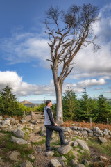 Adult men standing on a rock next to large wither tree and looking at view, Sugarloaf Mountain in background. Hiking in Wicklow Mountains, Ireland