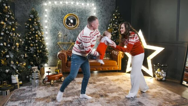 Cheerful mom and dad play with his newborn baby at Christmas night. Concept of family celebrate New Year at home, together winter holiday, decorated tree garland light, festive same costume
