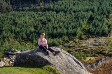 Naklejka na ściany i meble Young, smiling girl sitting on a rock, illuminated by sunlight with forest and valley in background. Hiking in Wicklow Mountains, Ireland