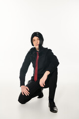 cosplay man in black clothes and wig with red tie posing on haunches looking at camera on white