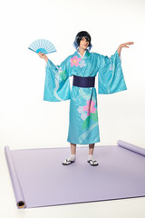 expressive man in blue kimono and wig posing with hand fan on purple carpet and white backdrop