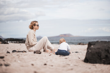 Mother enjoying winter beach vacations playing with his infant baby boy son on wild volcanic sandy beach on Lanzarote island, Canary Islands, Spain. Family travel and vacations concept. - obrazy, fototapety, plakaty