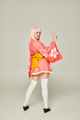 young woman in blonde wig and pink kimono with white knee socks showing victory gesture on grey