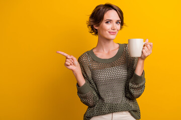 Photo of attractive young woman point empty space hold coffee mug wear trendy knitwear khaki clothes isolated on yellow color background - Powered by Adobe