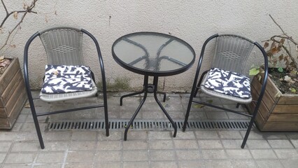 A metal table with a glass top and wicker chairs with cushions stand on a tiled area near a plastered wall.  Nearby are wooden tubs with withered ornamental plants