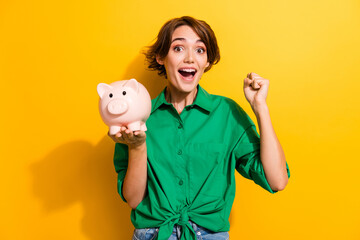 Photo of lucky excited girl dressed green shirt rising fist holding piggy cash box isolated yellow color background - obrazy, fototapety, plakaty