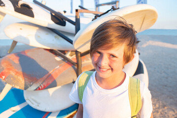 smiling child in a t-shirt with a backpack at the surf station on the beach in summer. joyful 10 year old child stands next to the windsurf boards, waiting for a training.	
