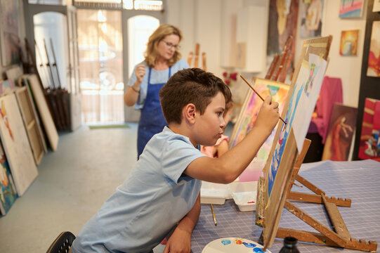 Authentic side portrait of a handsome preteen schoolboy artist focused on painting on easel in a creative art studio