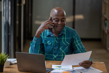 Black senior man working happily with document report. Using the phone talking with team.