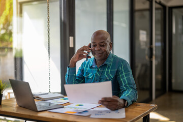 Black senior man working happily with document report. Using the phone talking with team.