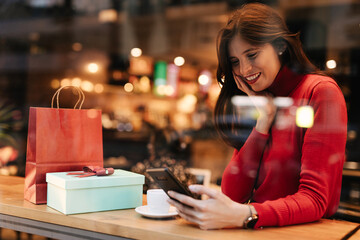 Young smiling woman using phone while drinking coffee in cafe.