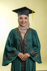 A female muslim lady wearing hijab and convocation attire smiling, isolated on yellow background
