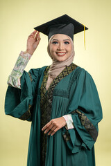 A female muslim lady wearing hijab and convocation attire smiling, isolated on yellow background
