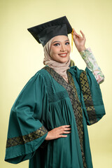 A female muslim lady wearing hijab and convocation attire smiling, isolated on yellow background