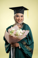A female muslim lady wearing hijab and convocation attire smiling, isolated on yellow background