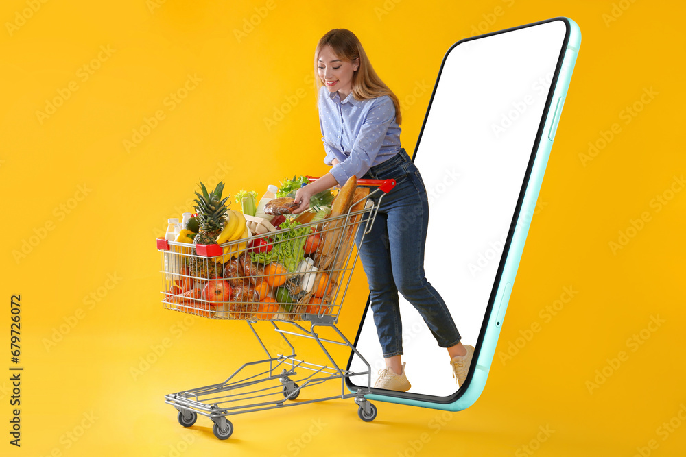 Poster Grocery shopping via internet. Happy woman with shopping cart full of products walking out of huge smartphone on orange background