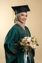 A female muslim lady wearing hijab and convocation attire smiling, isolated on brown background