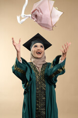 A female muslim lady wearing hijab and convocation attire smiling, isolated on brown background