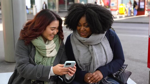 Young multiracial women having fun using mobile phone while waiting at bus station with city in the background during winter time - Friendship concept