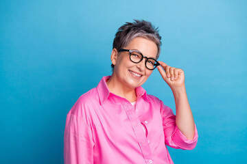 Photo of nice smart cheerful woman with short hair dressed pink shirt fingers touch glasses smiling isolated on blue color background