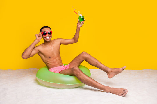 Full length photo of attractive excited shirtless man sitting inflatable circle rising cocktail glass isolated yellow color background