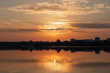 Scenic warm sunrise sun on cloudy sky reflecting in calm lake water. Peaceful morning in city park by the river