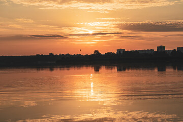 Scenic warm sunrise sun on cloudy sky reflecting in calm lake water. Golden morning in city park by the river
