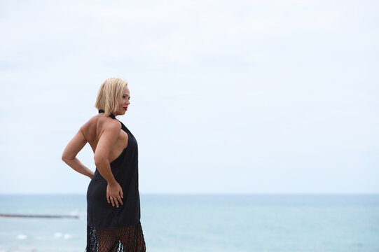 Mature, blonde and beautiful woman wears an elegant black dress and poses for the pictures happy and smiling while doing different poses. In the background the atlantic ocean in cadiz, spain.