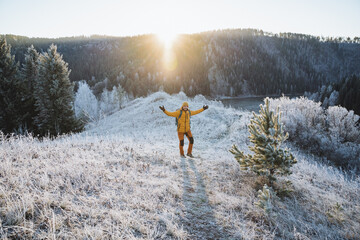 Naklejka na ściany i meble A joyful man stands in the mountains against the background of the glare of the sun, a beautiful day of one traveler, a winter hike in the mountains, harsh weather conditions.