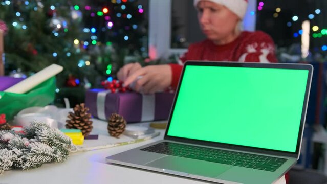Laptop with green screen on table against backdrop blurred of woman  in red sweater wrapping gifts boxes, glowing Christmas tree garlands, bokeh and xmas decoration, festive background New Year eve. 