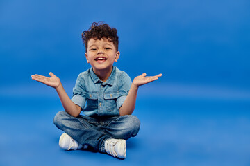 joyful and curly african american preschooler boy in denim clothes sitting and gesturing on blue