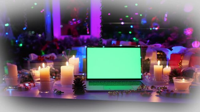 Portable laptop with chromakey on screen close up among xmas tree decor of burning candles, pine cones and flickering colored garlands, creating Christmas mood, during New Year time.
