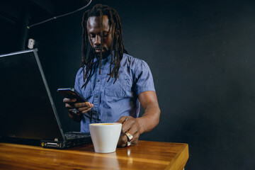 Freelance black man with dreadlocks inside cafe using phone working and holding cup of coffee.