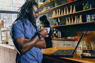 Happy freelance black man standing looking at laptop on restaurant counter, holding cup of coffee