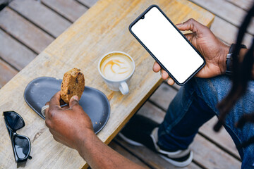 top view of unrecognizable young man holding phone and eating cookie with coffee, sitting outdoors