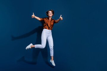 Full length photo of adorable confident lady wear brown shirt jumping showing two thumbs up empty space isolated blue color background