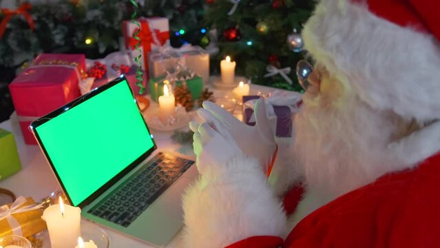 Santa Claus sitting with laptop green screen chroma key in cozy warm candlelight, among wrapped gifts, cones, fir branches, closeup side view. Xmas special offer. Happy New Year.