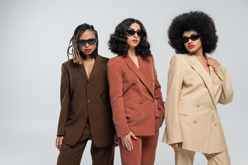 glamorous multiethnic girlfriends in colorful suits and dark sunglasses posing on grey, diversity