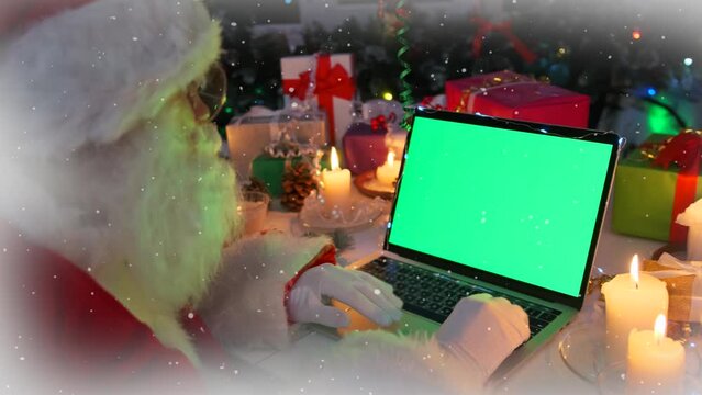 Modern Santa Claus at laptop with green screen, ordering gifts in online store, next to Christmas decor, present boxes, candles, cones, fir branches, glowing garlands, xmas tree, close up, side view.