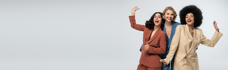 smiling multiracial girlfriends in stylish colorful suits waving hands on grey, horizontal banner