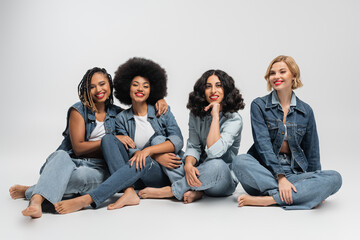 joyful and barefoot multicultural female models in blue jeans clothing sitting on grey backdrop