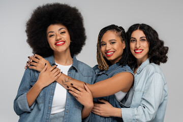 stylish and smiley multiethnic girlfriends in blue jeans wear looking at camera on grey, diversity