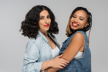 two carefree multiracial girlfriends in stylish denim attire smiling at camera on grey, togetherness