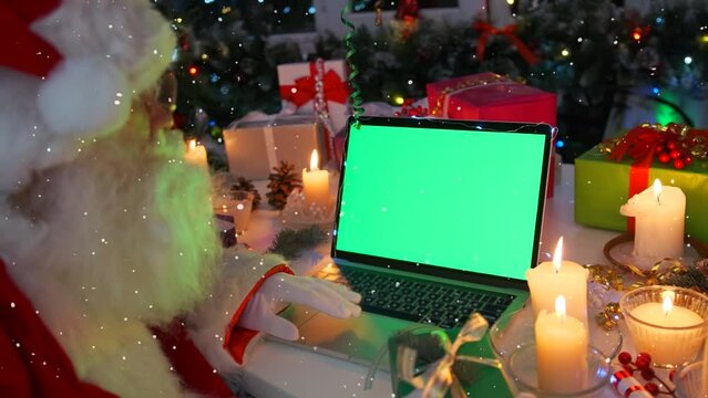 Santa at laptop with chromakey using touchpad on desk against glowing Christmas tree garlands, burning candles, gift boxes, pine cones and sparkling lights, festive background, New Year party event.