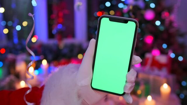 Mobile phone with chromakey screen in hand against backdrop of festive decorations of burning candles, Christmas gift boxes, New Year tree in garlands and tinsel. Space for social media holiday advert