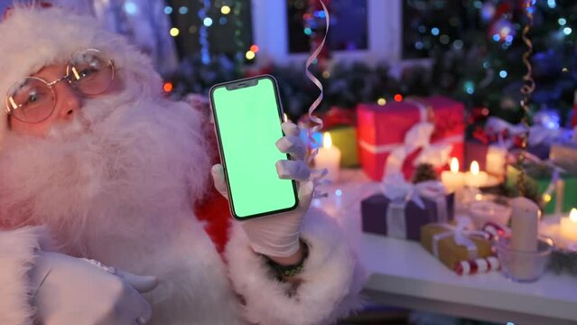 Bearded Santa in glasses holds out smartphone with green screen show place for advertising New Year sales, against of Christmas decorations of flickering bokeh lights, present gifts, Christmas tree