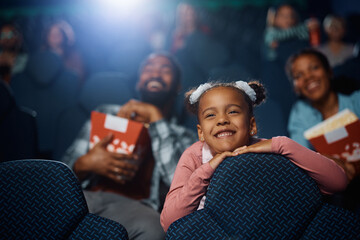 Smiling African American girl watching movie with her parents in cinema. - obrazy, fototapety, plakaty