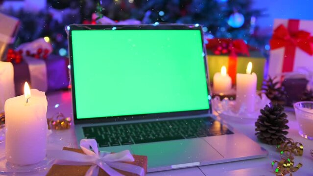 Static laptop with green chromatic screen close-up, beautiful xmas decor, burning candle, gift boxes, tinsel, bokeh and fir tree. Christmas background. New Year holidays concept.