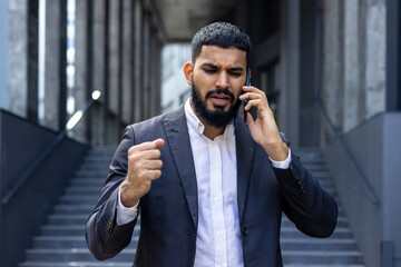 Upset and angry Indian male businessman standing outside an office center and talking on the phone emotionally - obrazy, fototapety, plakaty
